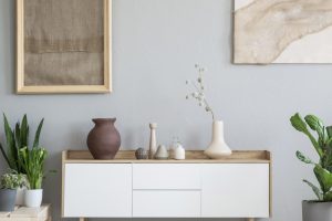 White cupboard between plants in grey living room interior with burlap in a frame and brown carpet. Real photo